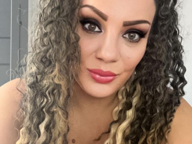 RachelI's curly gray and blonde hair, full pink lips, and dark makeup with thick eyebrows on a light brown face, looking at the camera with a slight smile.