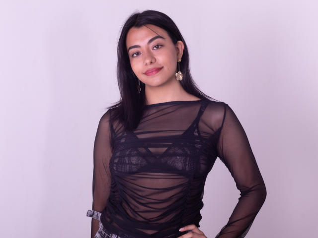 VeronicaJhones with black sheer top, showing her small boobs through lace bra, wearing gold earrings, standing with hand on hip, smiling softly, straight black hair, light pink background.