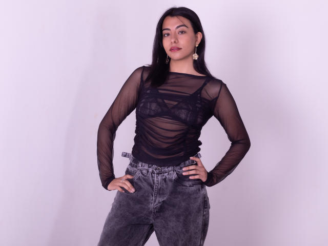VeronicaJhones with black sheer long-sleeve top showing her tits, wearing gray velvet pants, hands on hips, standing against white wall, long black hair, red lipstick, gold earrings.