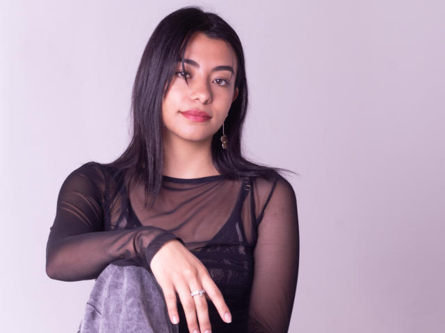 VeronicaJhones met een zwarte doorschijnende top, middelgrote borsten, bruine ogen, steil zwart haar, roze lippenstift, een ring aan de linkerhand, een grijze doek, een witte achtergrond, een subtiele glimlach, oorbellen, zacht licht..