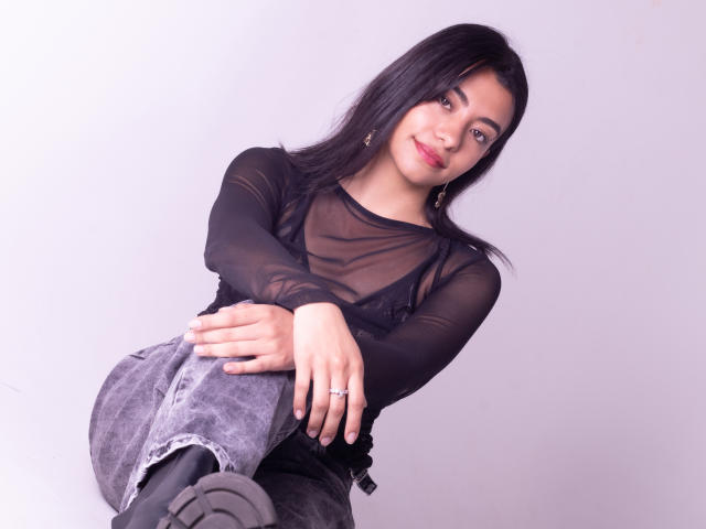 VeronicaJhones with black sheer top, showing her small tits, sitting with legs crossed, wearing grey jeans and black boots, smiling, ring on her finger, white background.