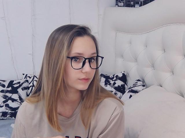 CaramellSex has blonde hair, black glasses, and a beige shirt, sitting on a white tufted bed with black-and-white pillows, looking at the camera with a slight smile.