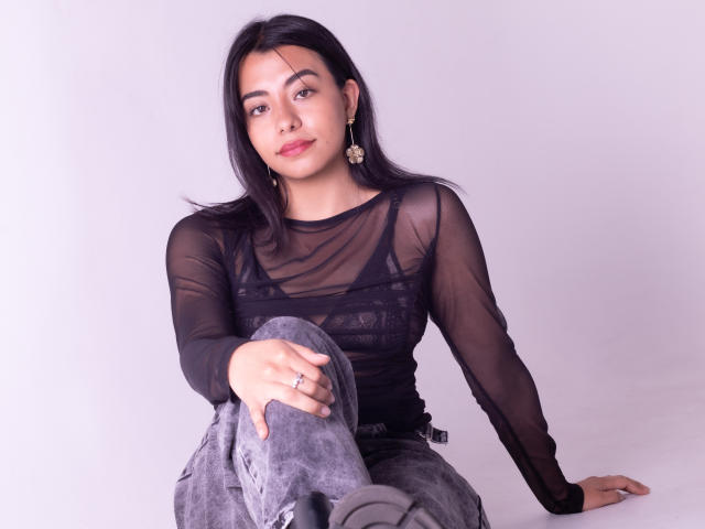 VeronicaJhones with black sheer top, showing tits, wearing gray jeans, sitting, black boots, earrings, ring, slight smile, medium skin, black hair, white background.