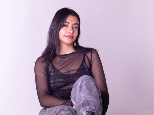 VeronicaJhones with long black hair, wearing a sheer black top showing her tits, gray velvet pants, and gold earrings, smiling softly.