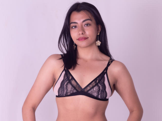 VeronicaJhones with black lace bra, pink underboob, small tits, light brown skin, black hair, earrings, slight smile, standing, looking at camera, arms at sides.
