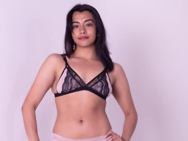 VeronicaJhones with dark hair, medium skin, wearing pink and black lace bra, showing her small tits, standing with hands on hips, smiling slightly.