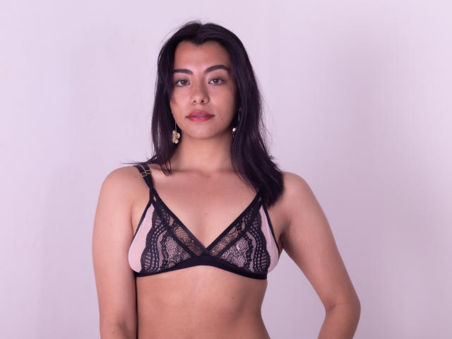 VeronicaJhones with medium brown skin, dark straight hair, wearing black lace bra, pink fabric, small to medium tits, standing, looking at camera, pink lipstick, gold earrings.