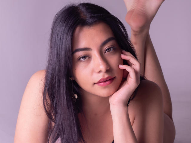 VeronicaJhones with long black hair, brown eyes, and pink lips, lying on her stomach, touching her cheek with one hand, legs up, showing her bare ass.