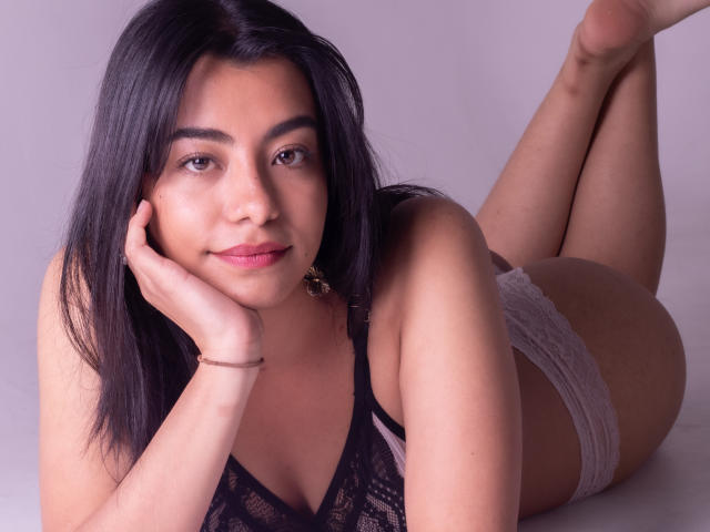VeronicaJhones with black lace bra, white lace panties, lying on her stomach, legs up, hand on cheek, dark hair, pink lipstick, earrings, bracelet, soft smile, cute, sexy.