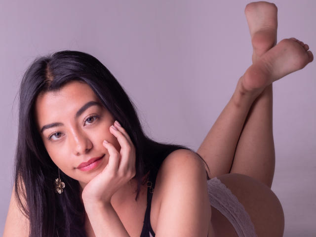 VeronicaJhones with long black hair, lying on her stomach, legs up, wearing white lace panties, black bra, hand on cheek, looking at camera, soft smile, bare soles.