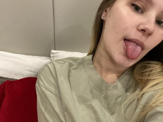 CaramellSex, blonde, tongue out, green shirt, small nose piercings, light skin, visible freckles, slightly messy hair, small earrings, red and white pillow in background.