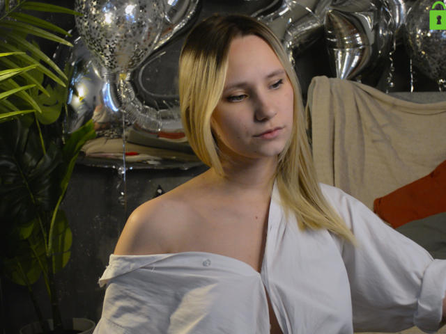 CaramellSex with blonde hair, white shirt slipping off her shoulder, exposing her collarbone, looking down with a slight smile, silver balloons in the background, green leaf on left.