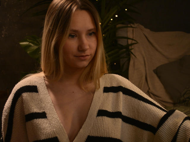 CaramellSex with blonde hair, wearing a white, black-striped, V-neck sweater, looking slightly to the side with a subtle smile. Her small tits are visible under the sweater.