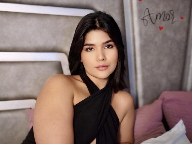 ScarlletZhen with medium brown skin, black hair, and brown eyes, wearing a black halter top, showing her bare shoulders and upper chest, sitting with a pink pillow.