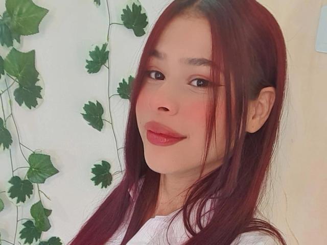 EuphoriaRush a les lèvres rouges, les cheveux châtain-roux et un sourire adorable. Elle porte une chemise blanche avec des lianes vertes en arrière-plan. Ses seins ne sont pas visibles..