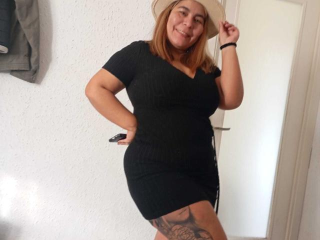 LouiseSexBigBoobs in a black rib-knit dress, holding a beige hat, smiling, showing a tattoo on her right thigh, standing with her hand on her hip.