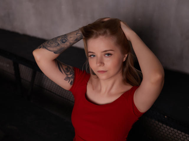 SirenaHardmann with hands in blonde hair, red shirt, tattooed arm, blue eyes, and slight smile, sitting against dark metal bench. Her tits are small, her pussy is not visible.