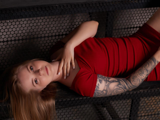 SirenaHardmann, with red dress, lying on black hexagonal-patterned surface, tattooed arm, hand on neck, brown hair, green eyes, looking at camera, soft light, relaxed, sexy.