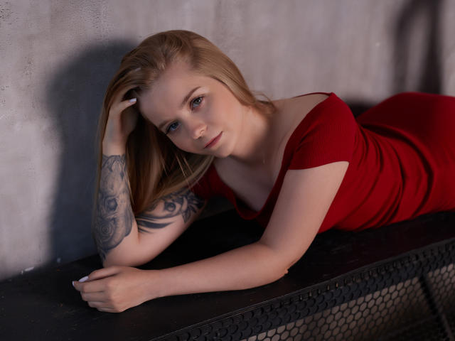 SirenaHardmann with blonde hair, red dress, tattooed arm, lying on black metal, leaning on hand, looking at camera, blue eyes, soft light, subtle makeup, sexy, relaxed.