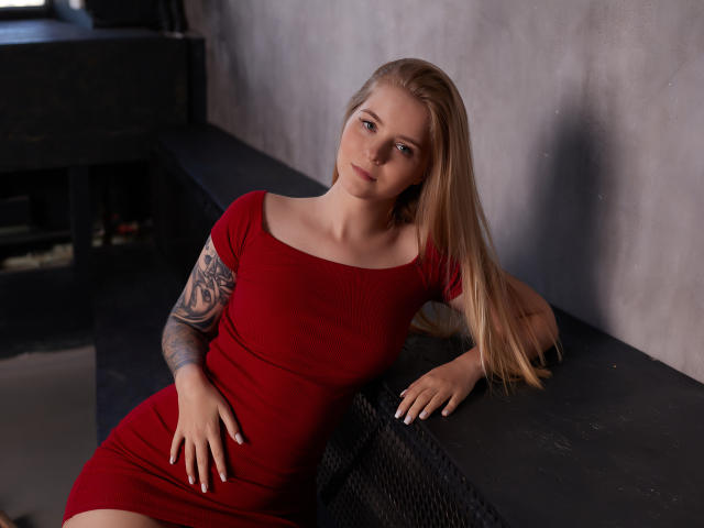 SirenaHardmann with long blonde hair, red dress, tattooed arm, and blue eyes, leaning on black bench, right hand on her ass, left arm resting on bench.