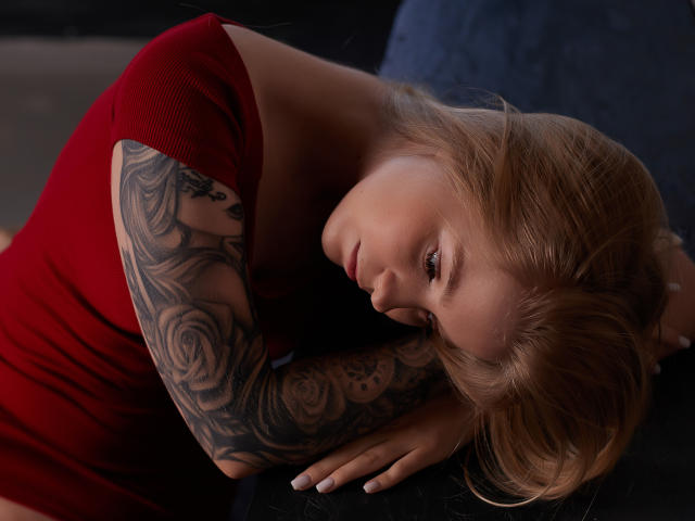 SirenaHardmann with blonde hair, red dress, tattooed arm, and pouty lips, lying down with head turned, eyes half-open, looking sexy and sultry. Boob, arm tattoo, dark background.