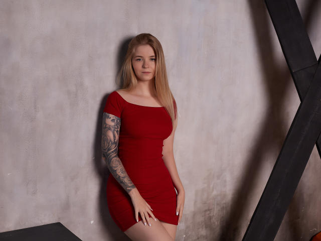 SirenaHardmann, blonde, red dress, tattooed arm, standing, hands on thighs, subtle smile, wall behind, black metal frame on right. Boobs, tits, ass, pussy, dress, tattoo.