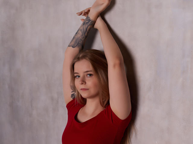 SirenaHardmann with arms raised, wearing a red shirt, has a tattoo on her right arm, and a slight smile on her face, standing against a textured wall.