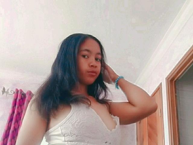 Brigitah with long black hair, wearing a white lace top, hand on head, looking at camera, small tits, blue bracelet, light brown skin, pink curtains in background.