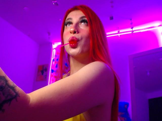 SammyViolet with red hair, pouty lips, red lollipop in mouth, tattoos on arm, under pink and purple neon lights, looking up, wearing yellow top, in selfie.