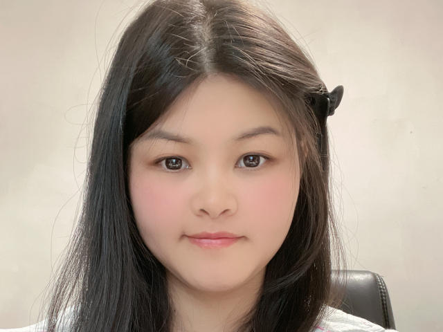 AdaCn has medium-length black hair with a black hair clip on the right side, brown eyes, pink cheeks, and a subtle smile. She's wearing a white top.