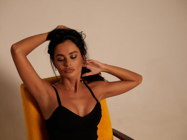 RinaNaash with arms raised, black tank top, yellow chair, brown skin, dark hair, closed eyes, subtle makeup, earrings, small nose ring, sexy, confident, soft lighting, relaxed pose.