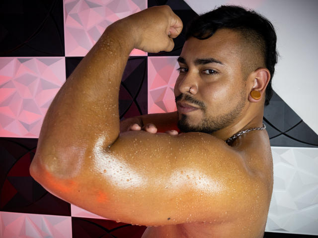 BrianMarly flexing a shiny, muscular arm with droplets of sweat, dark short hair, beard, gold ear plug, and chain necklace, looking at the camera with a slight smirk.