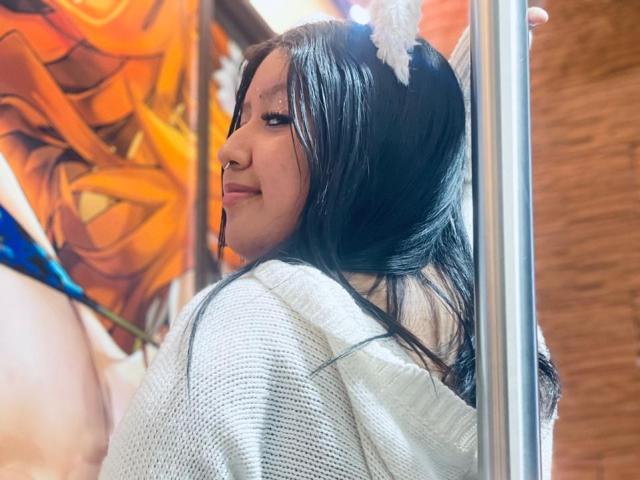 Daka69 with long black hair, white fuzzy headband, white knit sweater, looking over shoulder, smiling, standing next to a mirror, reflecting her upper body and boobs.