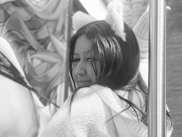 Zwart-wit foto van Daka69 met kattenoren, lang donker haar en een subtiele glimlach, die een gebreide trui draagt en over haar schouder kijkt, met een reflecterend oppervlak in de buurt..