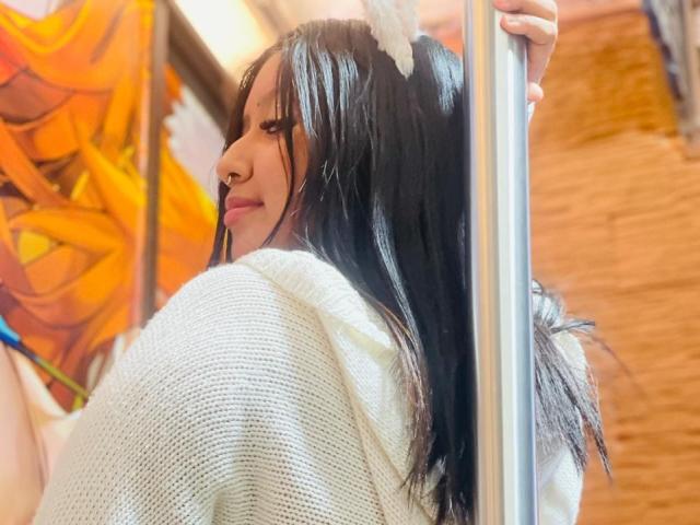 Daka69, com cabelo preto longo, suéter branco de tricô, piercing no nariz, segurando um mastro, sorrindo para a câmera, perfil direito, em ambiente interno. Sem seios visíveis, sem nádegas ou vulva visíveis..
