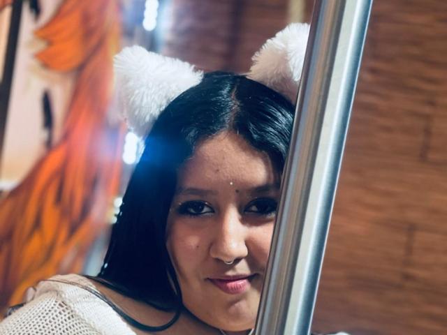 Daka69 with black hair, white cat ears, and nose ring, peering through a metal pole, wearing a white top, looking cute and slightly sexy.