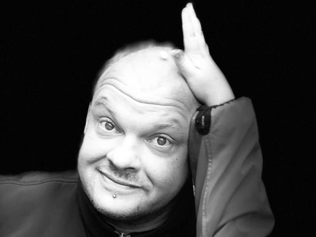 Magic-Mike-81-hot, bald with a small mustache, smiling, has a lip piercing, and is holding his head with one hand, wearing a jacket, in a black-and-white photo.
