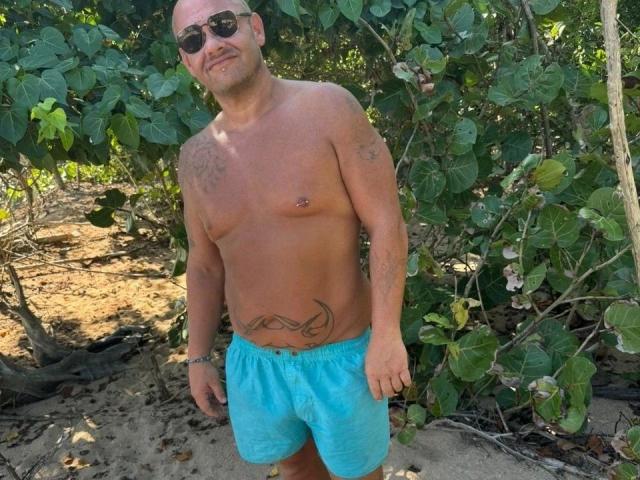 Magic-Mike-81-hot, shirtless, wearing blue shorts, sunglasses, tattoos on chest and belly, bald with beard, standing on sandy ground, green leafy background, nipple piercing visible, slight smile.