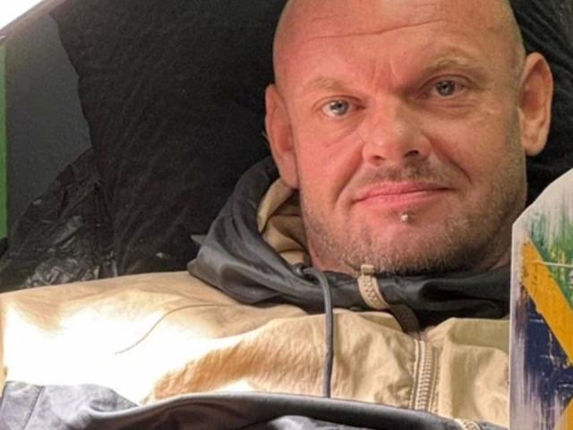 Magic-Mike-81-hot, bald with a light beard and nose piercing, wearing a beige and black hoodie, smiling slightly, is in a car with a magazine in the foreground.