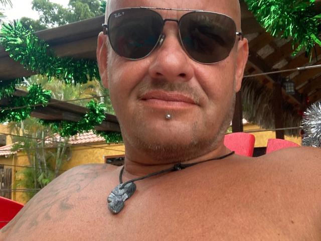Magic-Mike-81-hot with a bald head, tattoos, and sunglasses, sporting a lip piercing and a necklace with a gray stone, smiling at the camera.