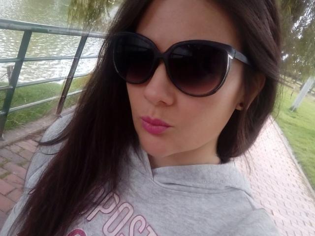 JakyKidman with dark brown hair, wearing black sunglasses, gray hoodie, and pink lipstick, pouting slightly, standing on a brick pathway near a green metal railing.