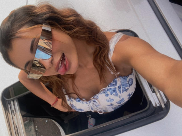 ArianaBellee with wavy brown hair, blue floral top, and reflective sunglasses, tongue out, sitting in a black car seat, wearing a red bracelet.