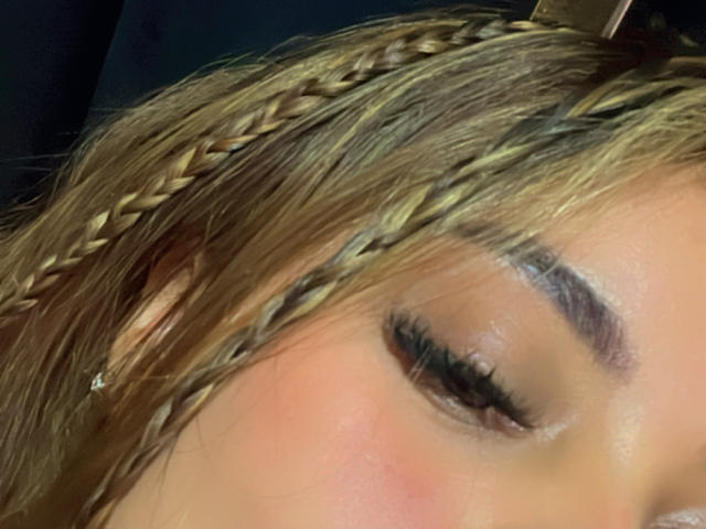 El rostro de Ariana Bellee, enfocándose en su cabello castaño trenzado, sus ojos ligeramente abiertos con largas pestañas y un ligero rubor rosado. Su ceja está cuidadosamente delineada. Se puede ver un pequeño arete..