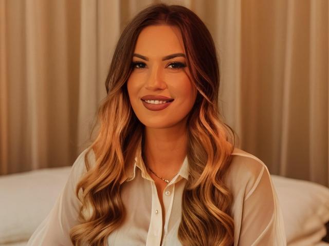 ZoeyMarie with long, wavy blonde hair, wearing a sheer white blouse, smiling softly, showing her tits slightly through the blouse, standing in front of beige curtains.