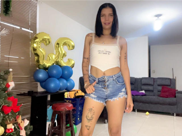 KaylinJulithe met zwart haar, een witte crop top, denim shorts, tatoeages, staand, blauwe ballonnen, een gouden ballon met het cijfer "21", een kerstboom en een grijze bank in een helder verlichte kamer..
