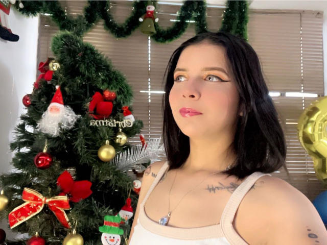KaylinJulithe, avec ses cheveux noirs, son rouge à lèvres rose, son débardeur blanc et ses tatouages au niveau du cou, se tient à côté d'un sapin de Noël décoré avec des ornements et une figurine de Père Noël..