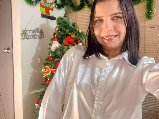 KaylinJulithe with shiny white button-up shirt, black shoulder-length hair, smiling with red lipstick, showing small white teeth, standing near decorated Christmas tree with red ornaments and green garland.