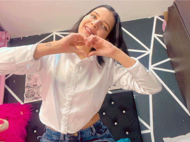 KaylinJulithe with black hair, smiling, wearing a white button-up shirt and blue jeans, making a heart shape with her hands, showing a tattoo on her right wrist.