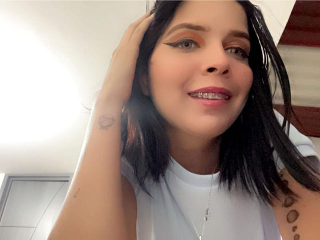 KaylinJulithe, avec ses cheveux noirs, sa chemise blanche et son rouge à lèvres rose, souriant en montrant ses dents, une main dans les cheveux, un petit tatouage en forme de cœur sur le bras, portant du khôl, dans une pièce bien éclairée..