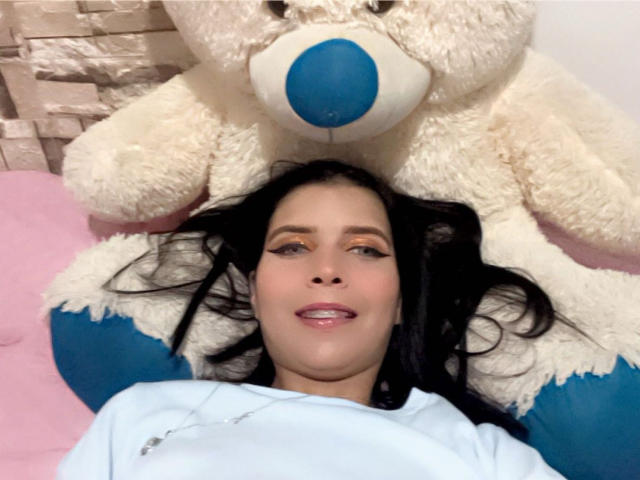 KaylinJulithe ligt op een roze bed, met zwart haar, een wit shirt, en een grote knuffelbeer erboven. De beer heeft een blauwe neus en ze glimlacht, terwijl ze omhoog kijkt. Ze draagt subtiele make-up en roze lippenstift. Ze ziet er schattig en ontspannen uit..